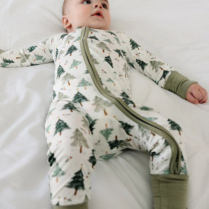 Angled view of baby model wearing Kidsely evergreen Christmas tree print bamboo viscose zipper sleeper on white bed sheet