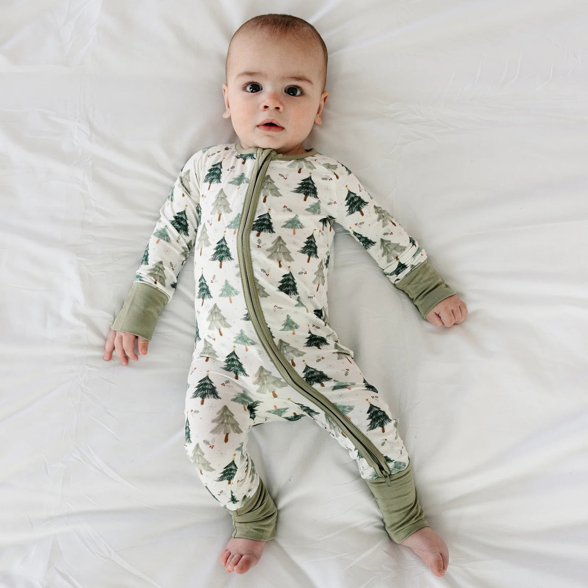 Baby model lying on white sheet wearing Kidsely evergreen Christmas tree print bamboo viscose zipper romper