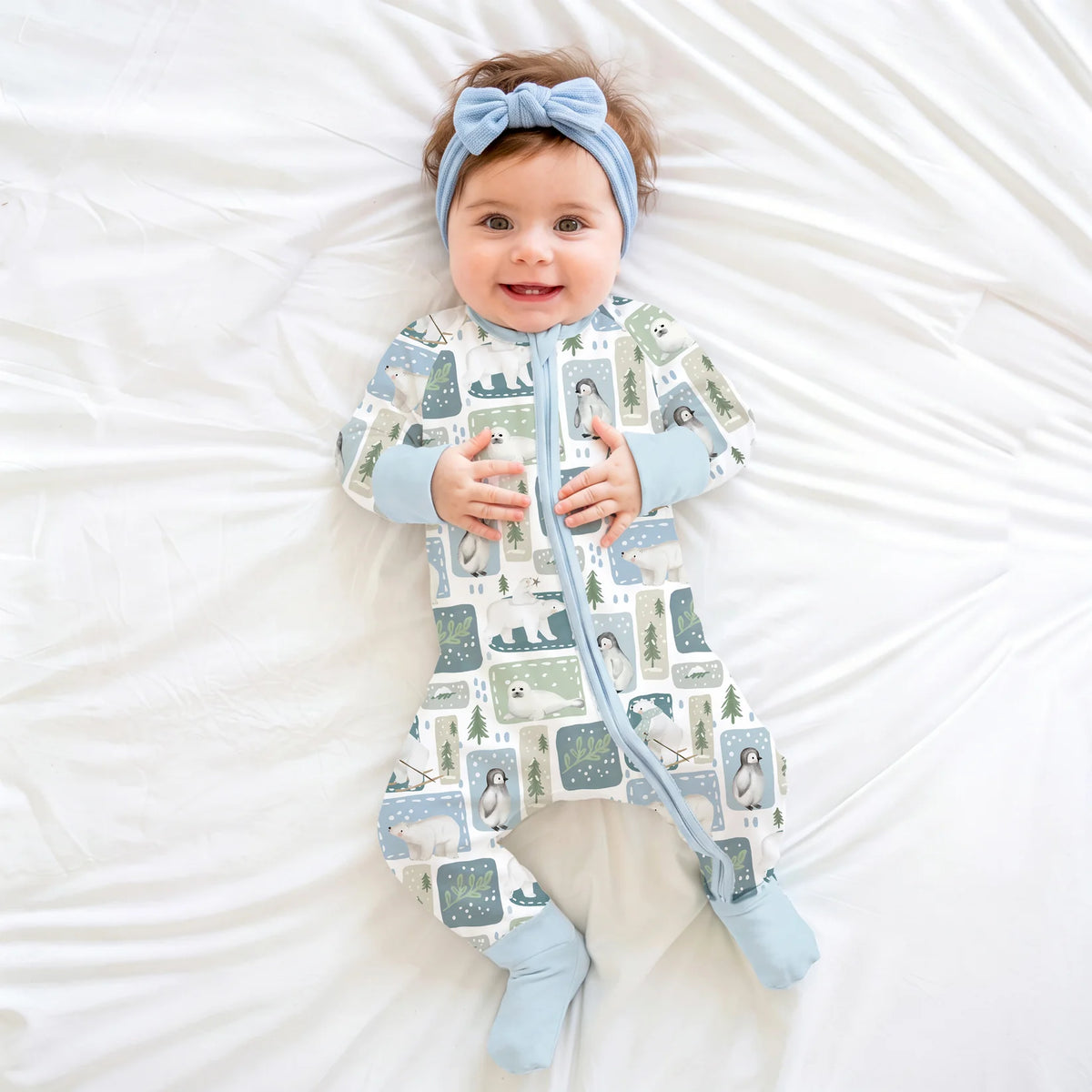 Baby model wearing blue headband and Kidsely winter polar bear and penguin print bamboo viscose zipper sleeper on white sheet