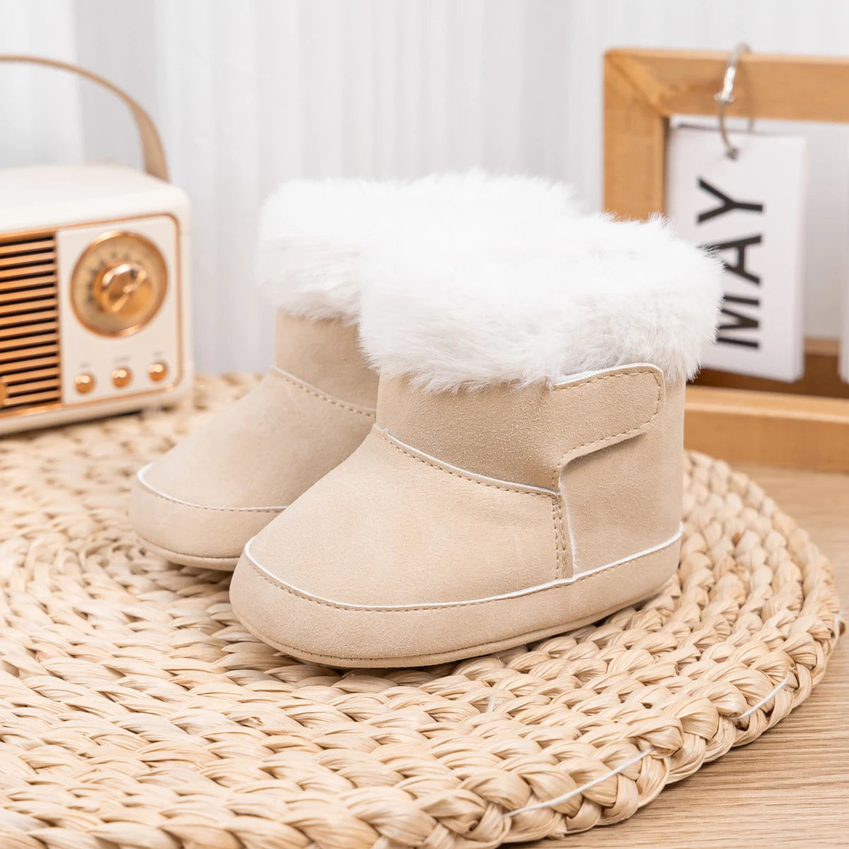 Beige baby soft sole crib boots displayed on a woven mat next to a vintage radio prop.