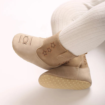 A baby wearing beige soft sole western cowboy booties with white tights, legs crossed to show toe embroidery details.