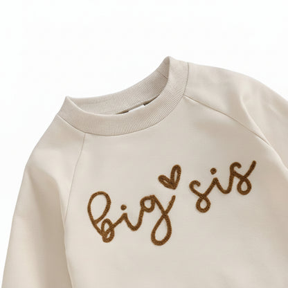 Close-up detail of the ribbed crewneck and brown script text reading Big Sis with a heart on the cream sweatshirt.