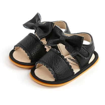 Pair of classic black leather baby girl sandals featuring a large decorative bowknot, scalloped edge band, and contrasting tan rubber soles.