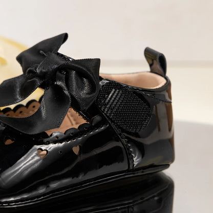 Side view of a black glossy baby girl Mary Jane shoe highlighting the secure hook-and-loop closure strap and scalloped edges.