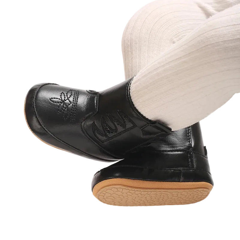 Close up of a baby wearing classic black western booties and white tights, with legs crossed to show the textured anti-slip rubber outsoles.