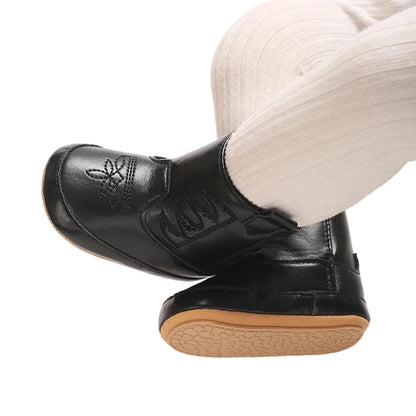 Close up of a baby wearing classic black western booties and white tights, with legs crossed to show the textured anti-slip rubber outsoles.