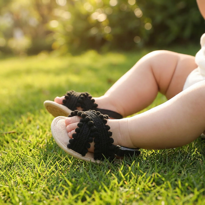 Baby girl wearing black pom pom sandals while sitting on green grass in the sun.