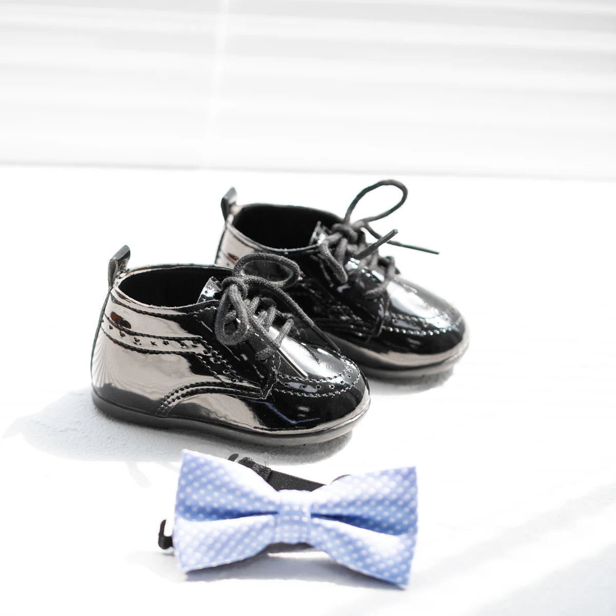Wider angled view of a pair of shiny black Kidsely patent faux leather baby formal brogue oxfords displayed with a formal light blue infant bowtie.