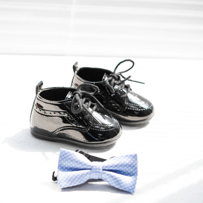 Wider angled view of a pair of shiny black Kidsely patent faux leather baby formal brogue oxfords displayed with a formal light blue infant bowtie.