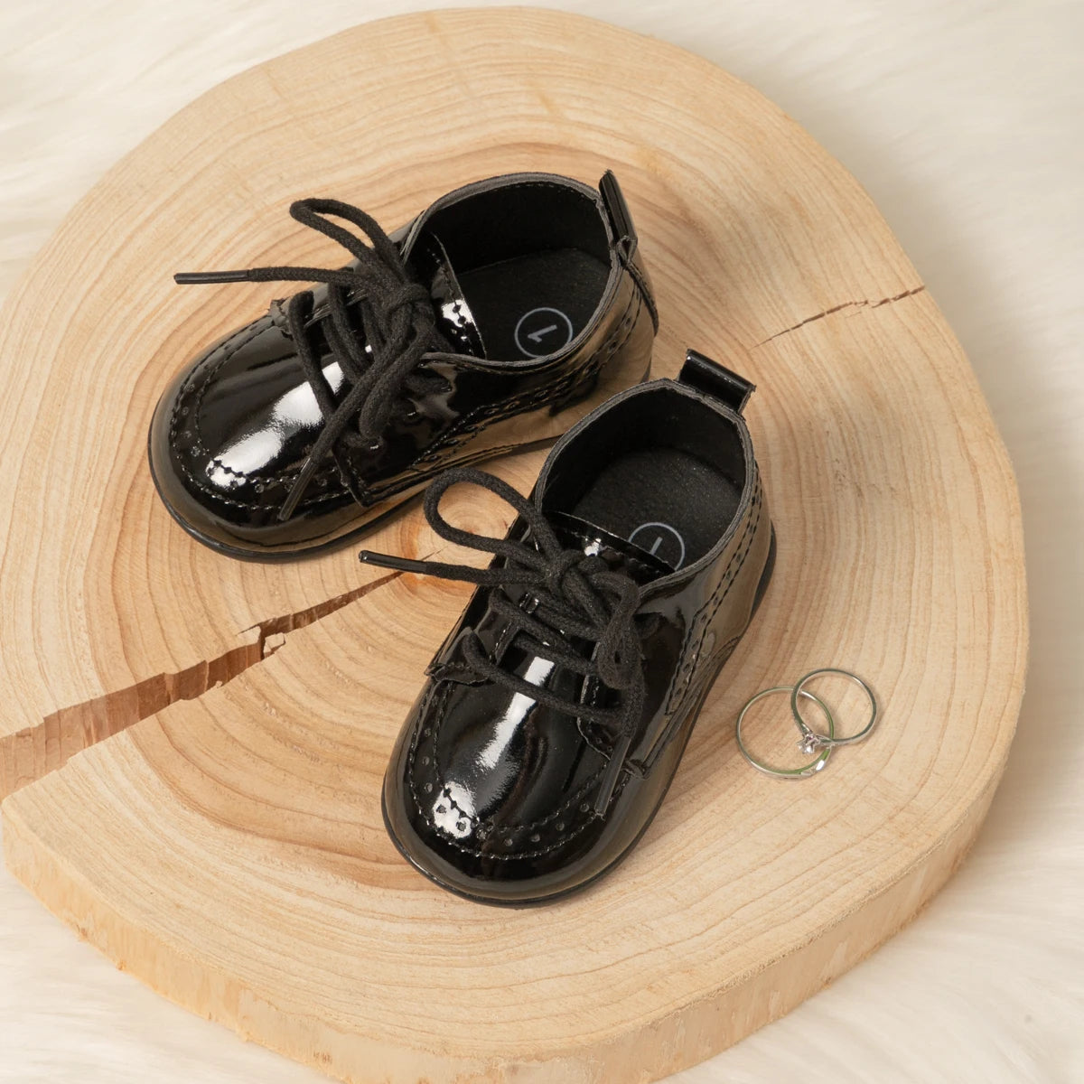 Flat lay view of a pair of shiny black Kidsely patent leather infant dress shoes styled beautifully on a rustic round wooden decorative slab.