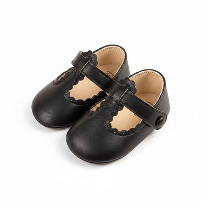 Pair of black PU leather baby girl T-strap crib shoes with scalloped edges.