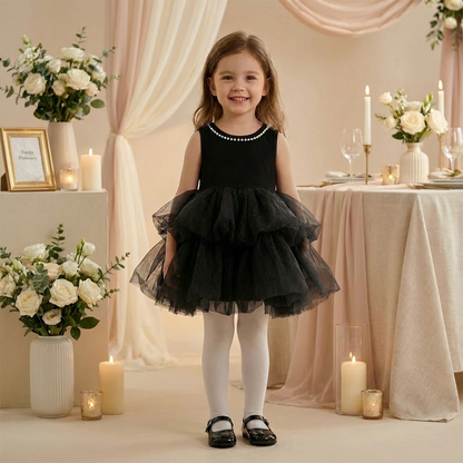 A young girl with long brown wavy hair, wearing the black tulle dress, smiles confidently in a warmly lit lifestyle room with white floral arrangements and candles.
