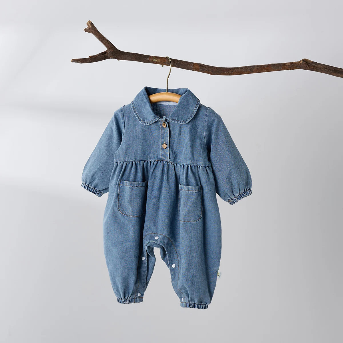 Full front view of a girls' blue denim long-sleeve jumpsuit hanging from a decorative wooden branch against a white wall.