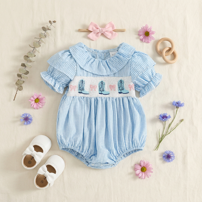 Flat lay of the blue gingham cowgirl baby romper styled with baby shoes, a pink headband, and floral accents.