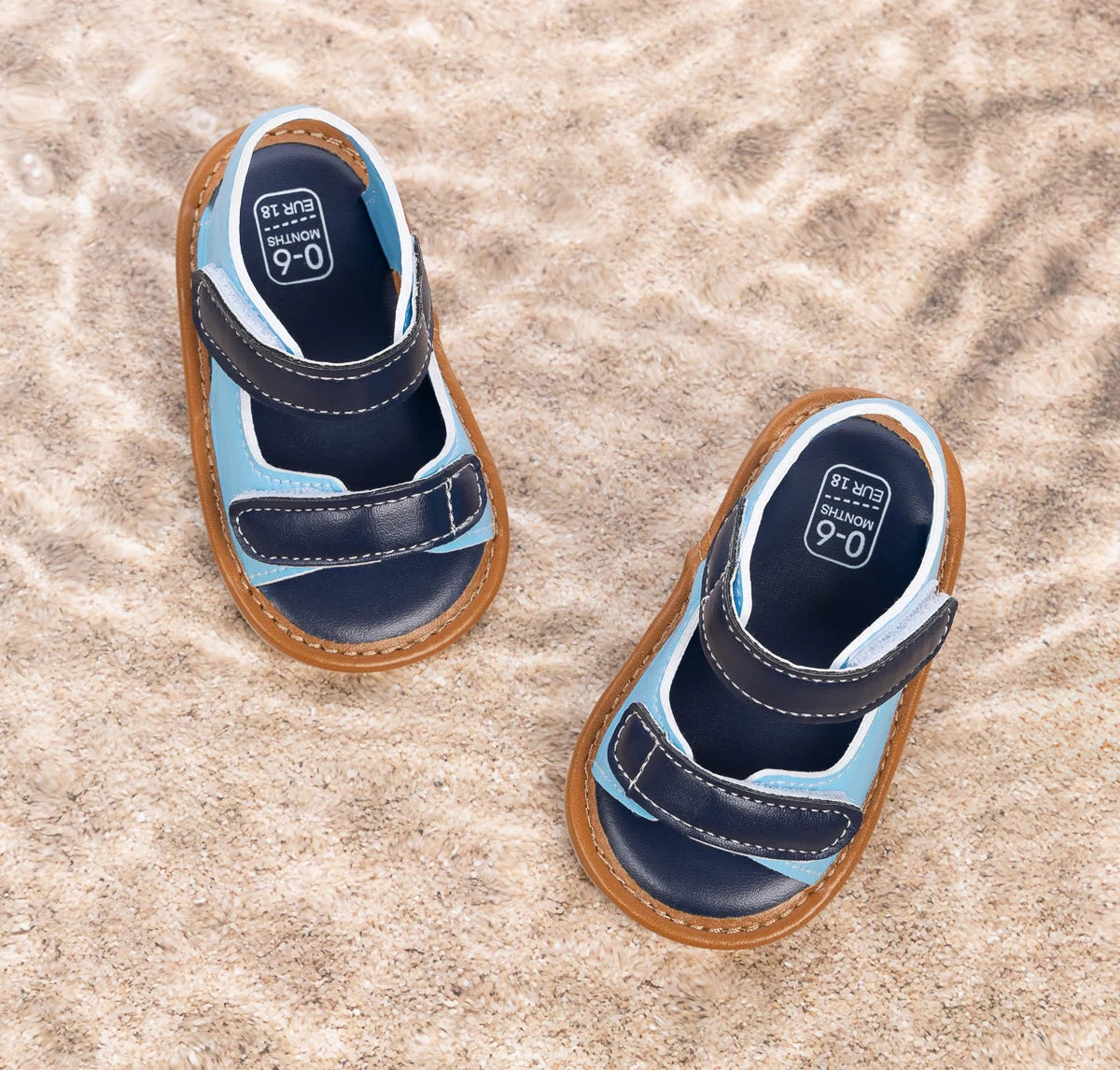 Overhead view of light blue and navy infant summer sandals resting on sand.