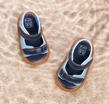 Overhead view of light blue and navy infant summer sandals resting on sand.