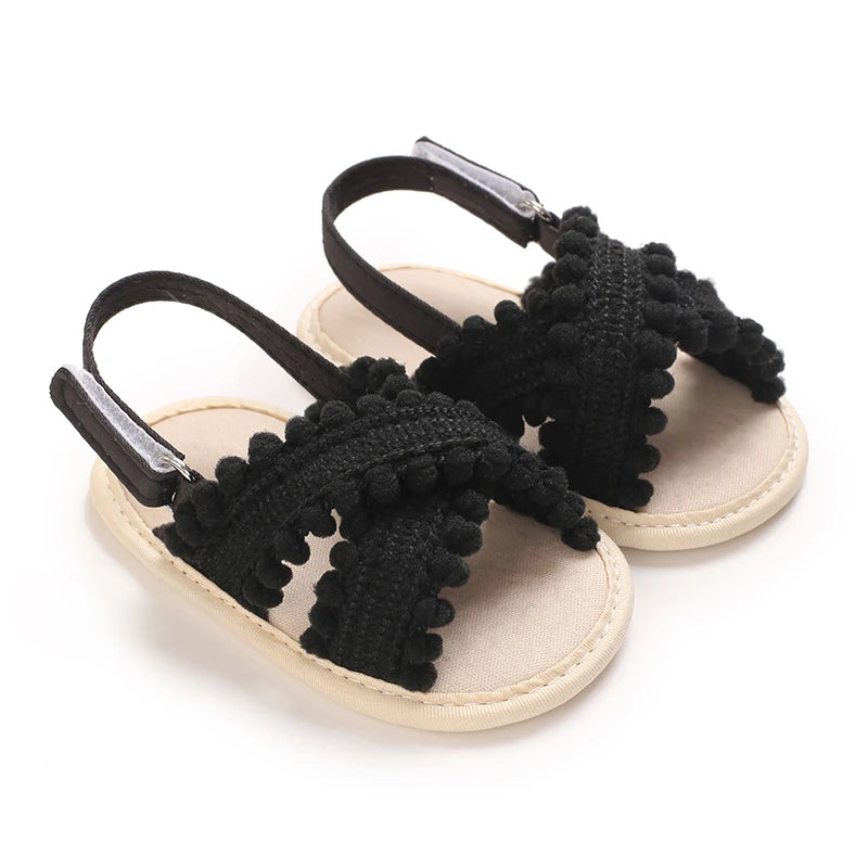 Black boho baby girl sandals with decorative pom poms and adjustable slingback strap.