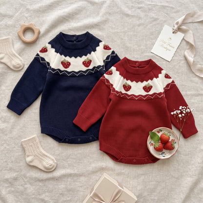 An overhead flat lay photograph of two cozy, knit baby sweater onesie rompers, one navy blue and one Christmas red, displayed on a textured beige linen background. Both rompers feature a charming cream-colored knit yoke across the shoulders with small knitted strawberry motifs. They are surrounded by matching cream baby socks, a wooden star-shaped teether toy, a small ceramic plate with fresh strawberries, delicate white flowers, and a gift box. 