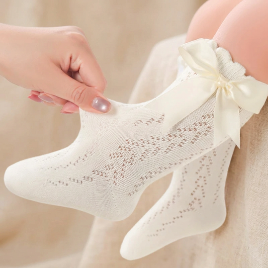 Close up of a white baby girl knee-high sock being gently stretched on a leg, showing the breathable cotton mesh pattern and satin bow.
