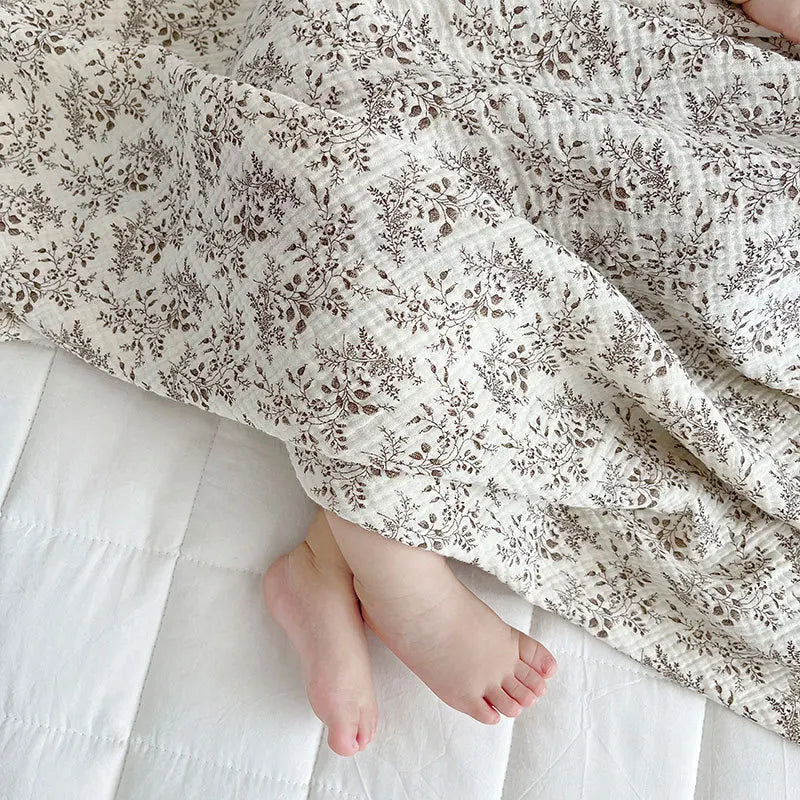 Baby's feet peeking out from beneath a lightweight, breathable taupe botanical print cotton muslin summer blanket.