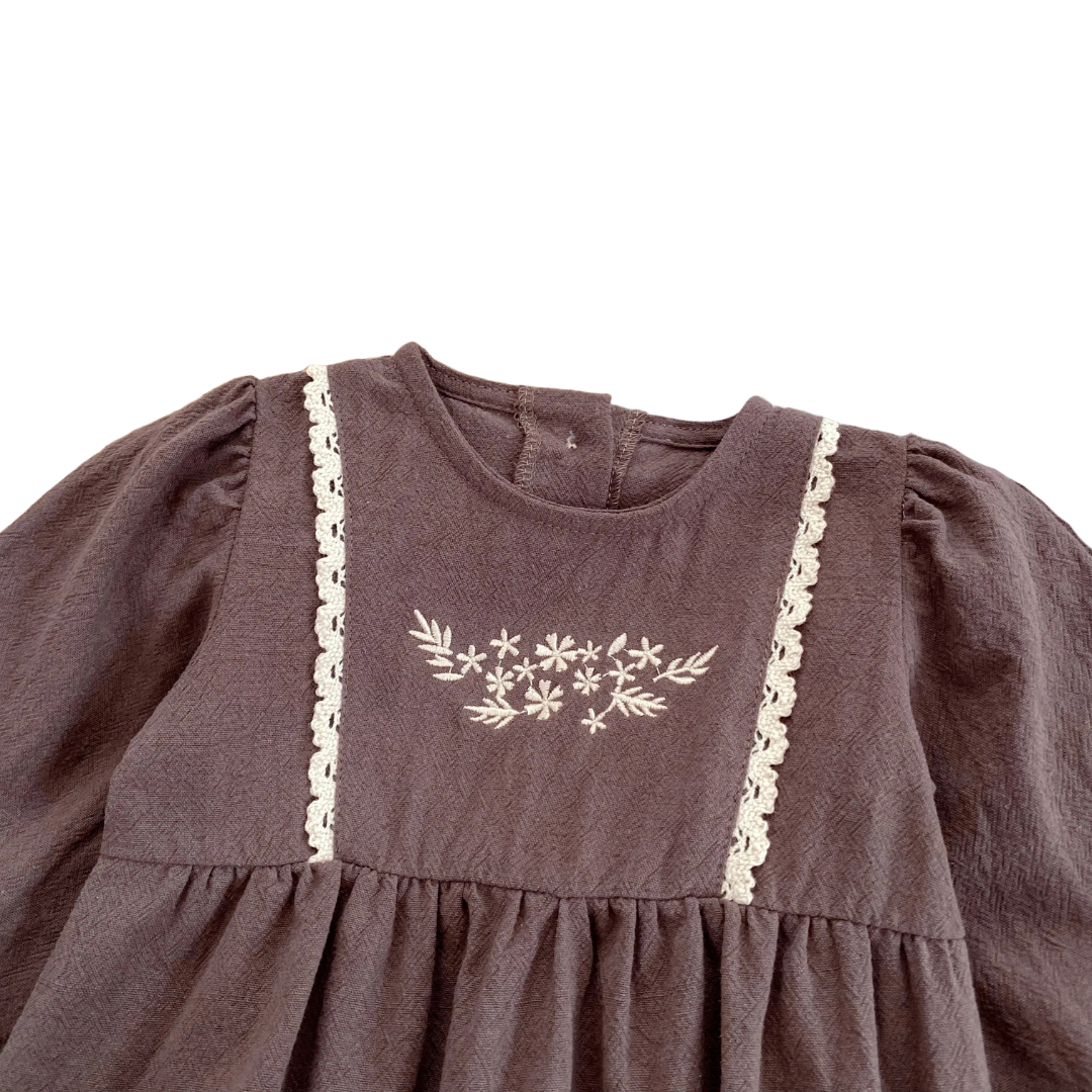 Close-up of the delicate white floral embroidery and vertical lace trim on the chest of the brown cotton baby girl bubble romper.