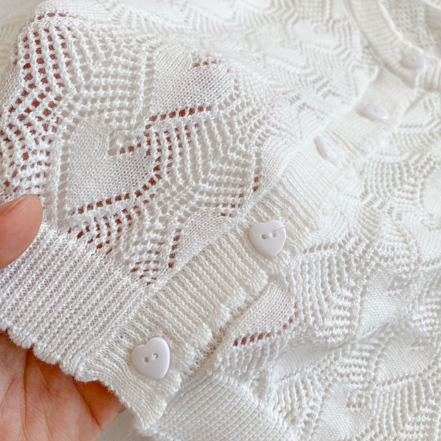 Close-up of the white baby girl cardigan showing the woven pointelle heart pattern and heart-shaped front buttons.
