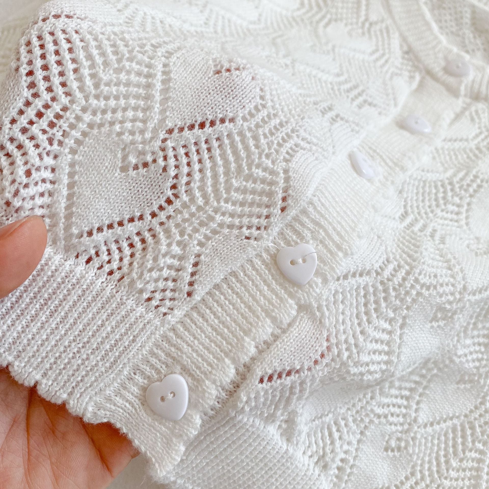 Close-up of the white baby girl cardigan showing the woven pointelle heart pattern and heart-shaped front buttons.