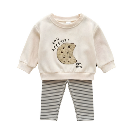 A two-piece baby outfit on a white background, featuring a cream ribbed long-sleeve pullover with a boucle cookie embroidery and "bon appétit!" text, paired with horizontal striped leggings.