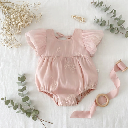 Flat lay of a pink embroidered bubble romper on a white surface decorated with dried eucalyptus, baby's breath, and a wooden teething ring.