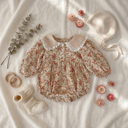 Flat lay style image of the cream vintage floral baby girl bubble romper styled aesthetically on a textured blanket with a baby bonnet and floral props.