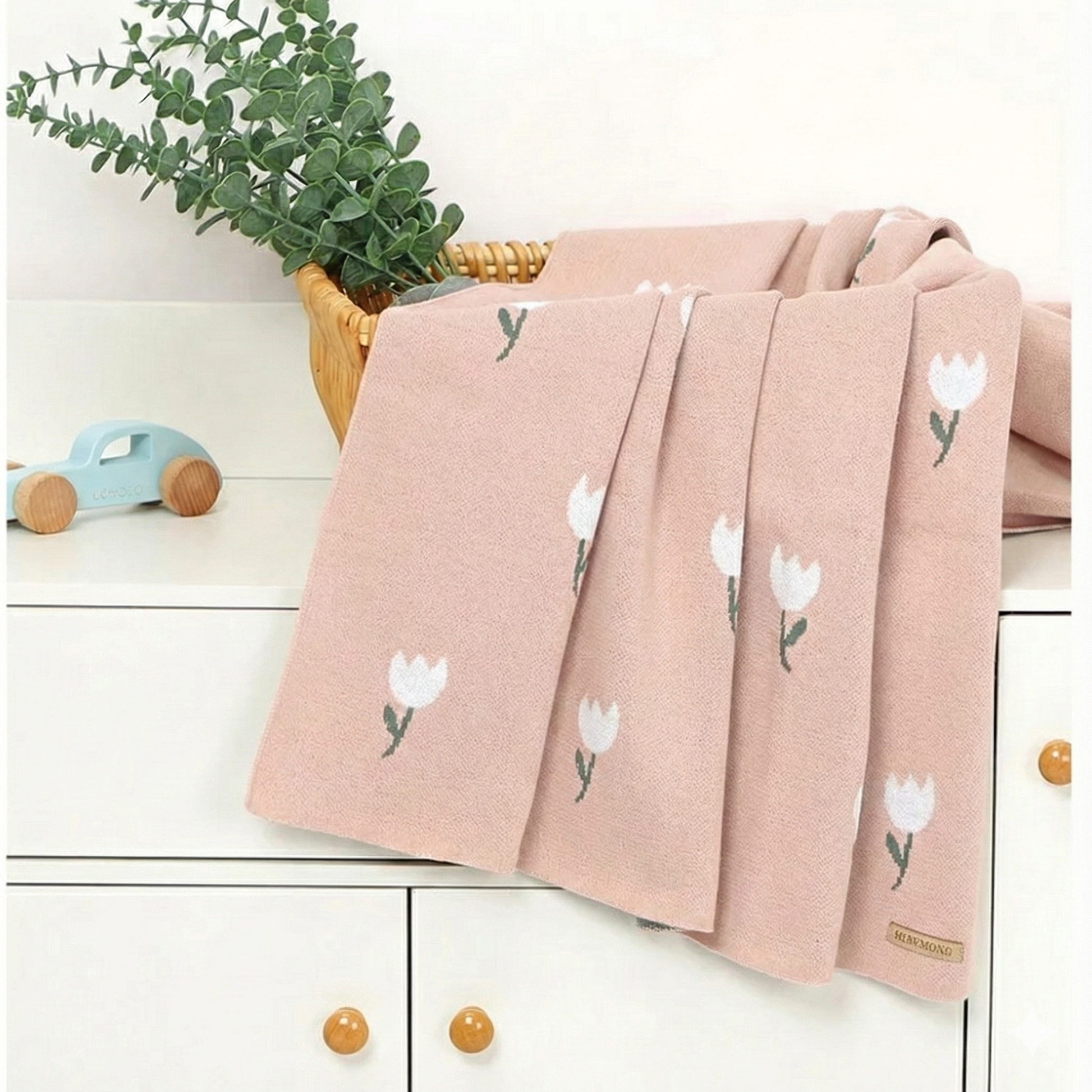 A blush pink cotton knit baby blanket draped elegantly over a white nursery changing table next to green foliage.