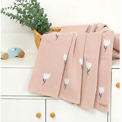 A blush pink cotton knit baby blanket draped elegantly over a white nursery changing table next to green foliage.