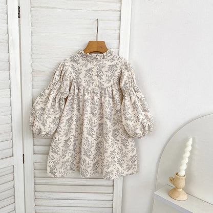 The back view of a vintage-inspired girls cream and gray floral dress hanging on a wooden hanger against a white slatted background.