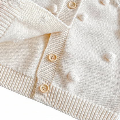 Close up of the cream baby cardigan showing the soft cotton knit, wooden-style buttons, and 3D pom-pom details.