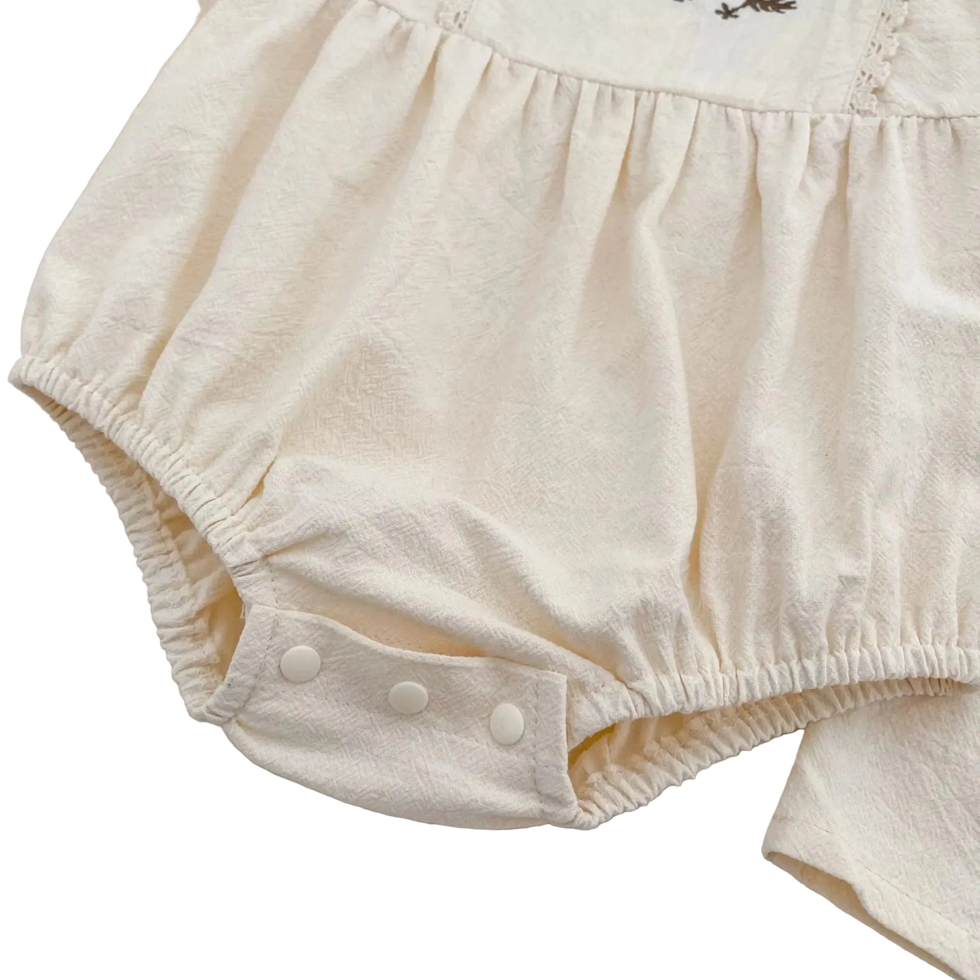 Close-up of the secure, fastened white bottom snap closures and gathered elastic on the cream infant bubble romper.