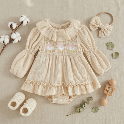Flat lay of a cream goose baby romper outfit styled with a matching headband, white baby booties, and a wooden rattle on a neutral background.