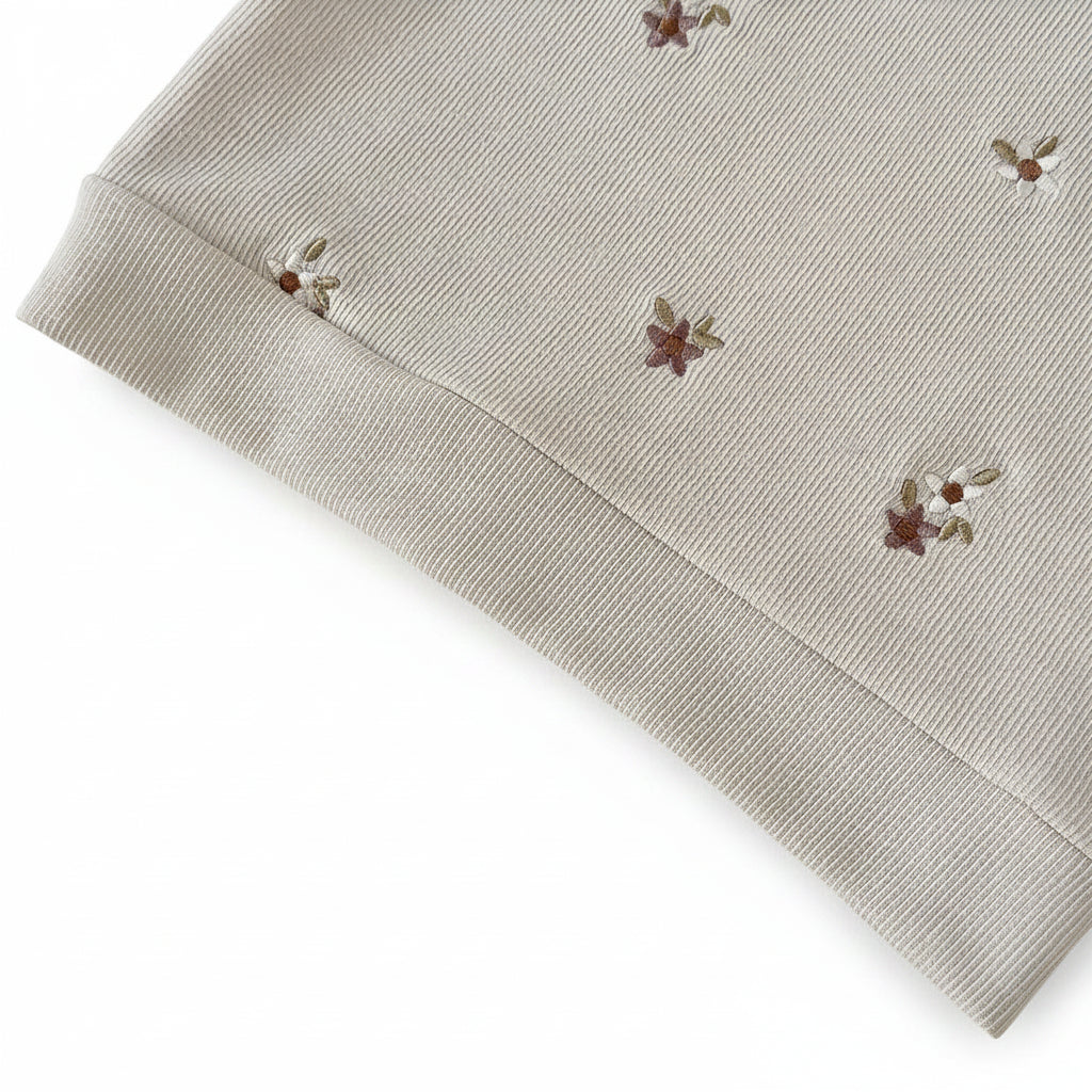 Close-up of the banded hem on a cream ribbed knit baby sweatshirt featuring earthy floral embroidery.