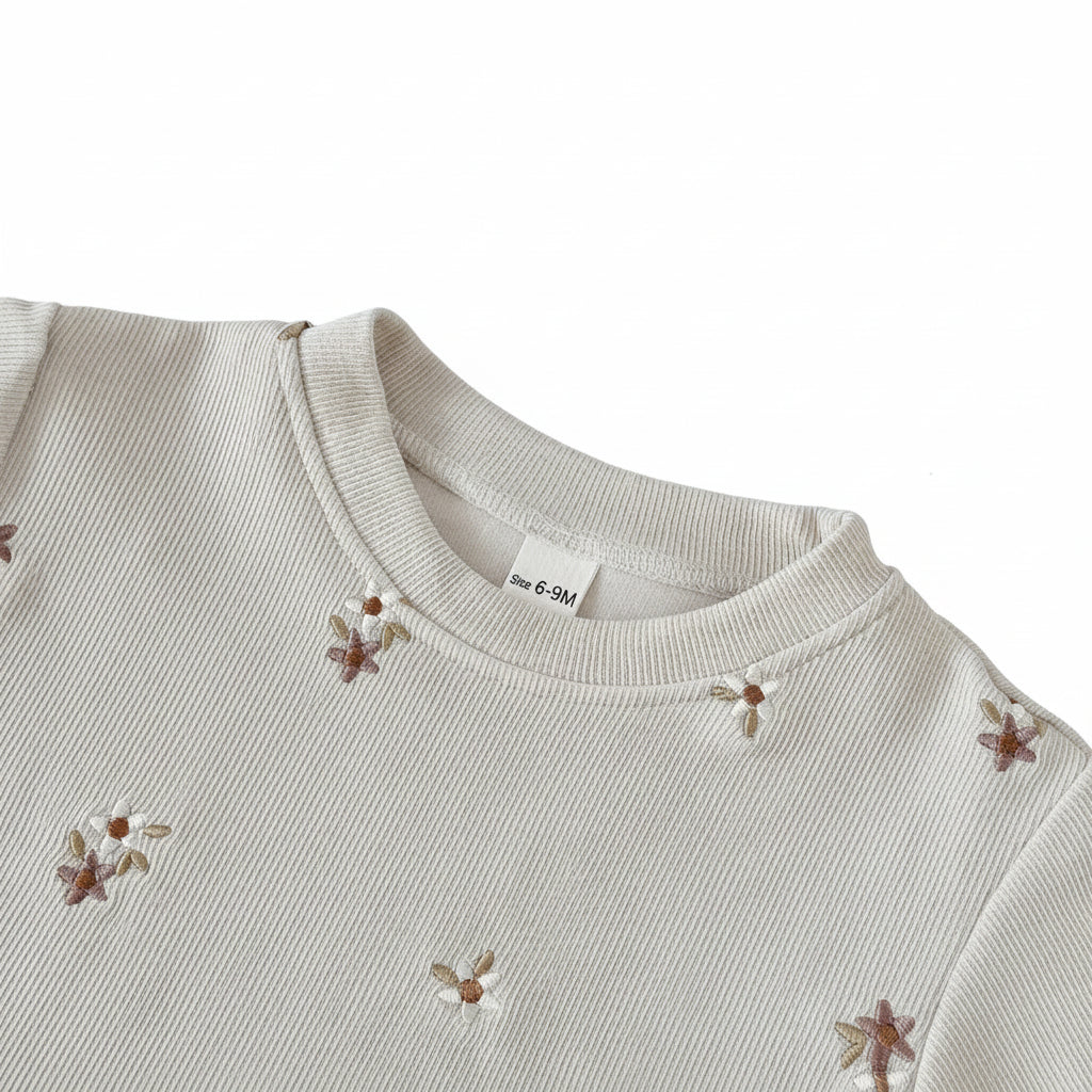 Close-up of a cream ribbed baby sweatshirt showing a banded crew neckline and delicate brown and white embroidered daisies.