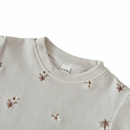 Close-up of a cream ribbed baby sweatshirt showing a banded crew neckline and delicate brown and white embroidered daisies.