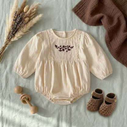Flat lay aesthetic photo of the cream floral embroidered baby girl bubble romper styled with brown baby booties, a wooden teething ring, and dried florals.
