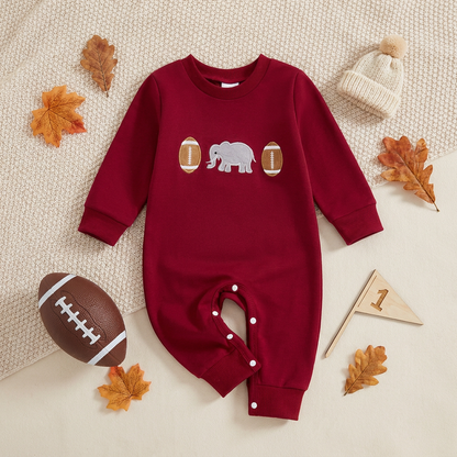 Styled fall flat lay of a premium crimson red infant long sleeve jumpsuit featuring vintage elephant and football chest embroidery, surrounded by autumn leaves, a mini football, a wooden pennant, and a baby beanie.