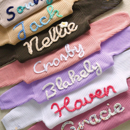 Stack of personalized knit baby sweaters showing custom hand-embroidered names in various font styles and yarn colors