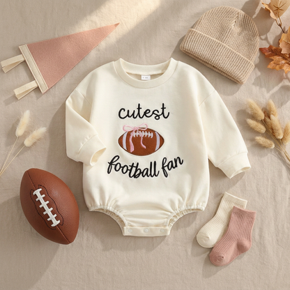 Flat lay of the cream "Cutest Football Fan" baby romper styled with a mini football, a pink pennant, a baby beanie, and cozy socks.