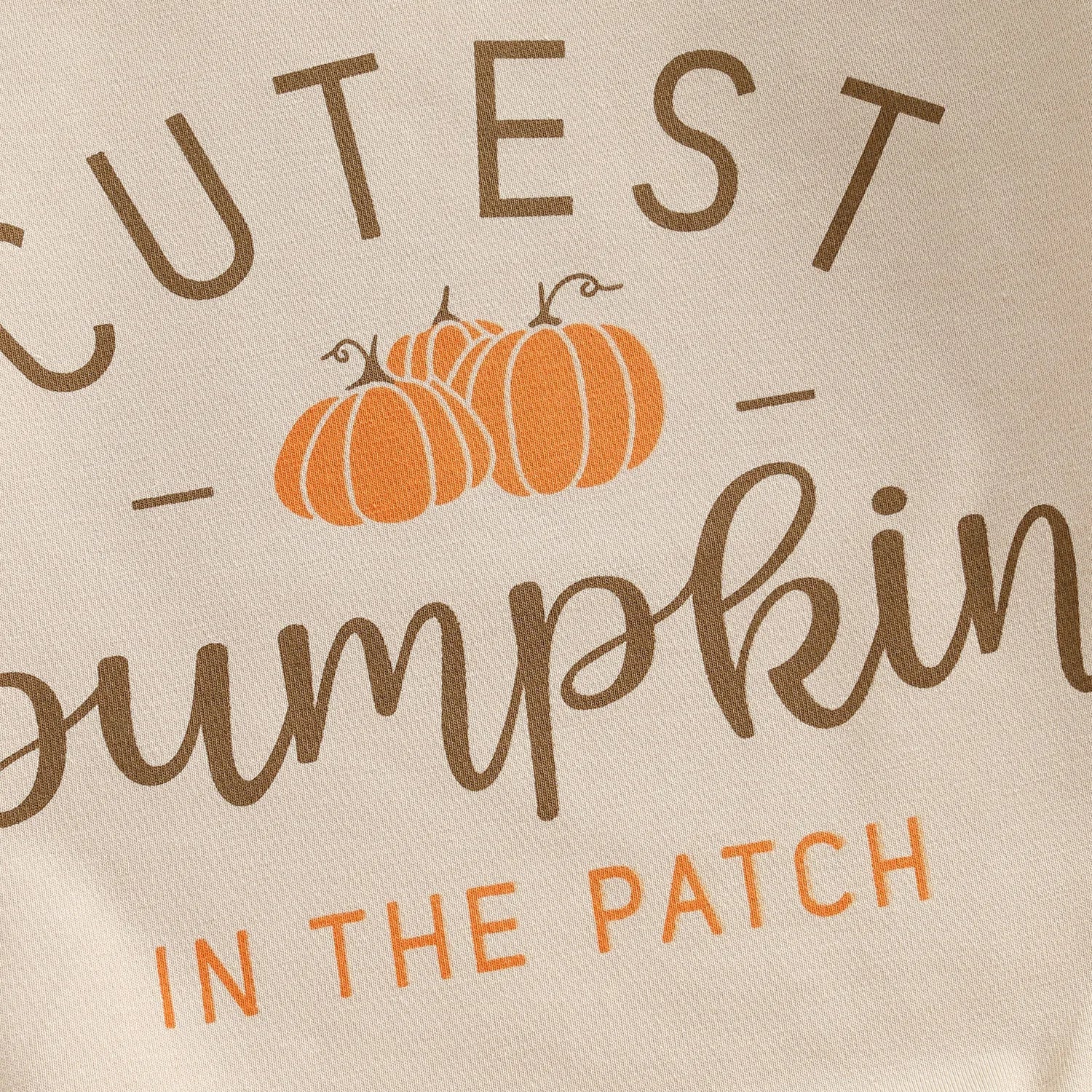 Close-up of the chest graphic on the beige romper reading Cutest Pumpkin in the Patch with three orange pumpkins.