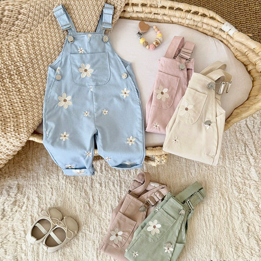 A styled collection of Kidsely toddler daisy overalls in light blue, blush pink, sage green, and cream, arranged in a woven basket with baby shoes.