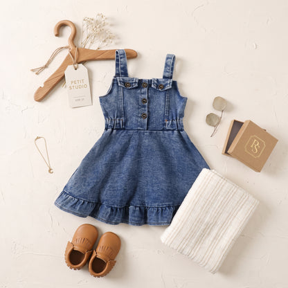 Flat lay outfit featuring the girls blue denim ruffle overall dress styled with brown baby shoes, a wooden hanger, and a white blanket.