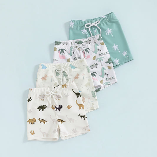 Toddler and baby boys quick-dry swim trunks in a variety of dinosaur and tropical palm tree prints, featuring ivory, white, and teal green colors with a decorative drawstring waist.