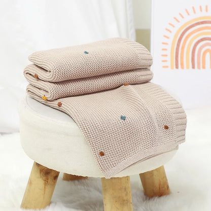 A Toasted Almond 100% cotton knitted baby blanket folded and draped over a modern white stool with wooden legs in a baby nursery setting.