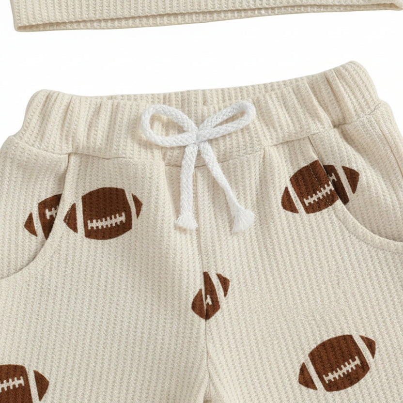 Close-up of the beige waffle-knit toddler pants showing the elastic waistband, white decorative drawstring, side pockets, and brown football print.
