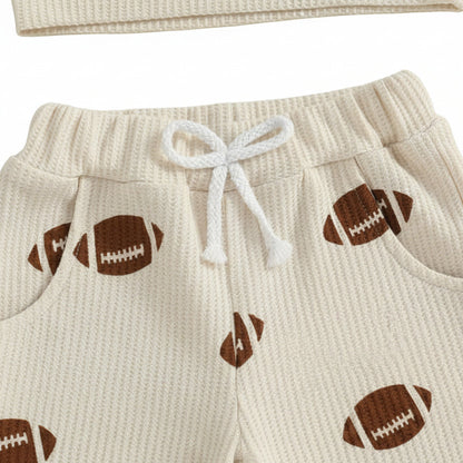 Close-up of the beige waffle-knit toddler pants showing the elastic waistband, white decorative drawstring, side pockets, and brown football print.